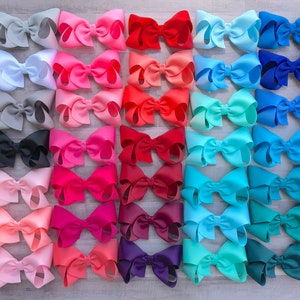 Pode incluir: Uma coleção de laços de cabelo grandes e coloridos em várias tonalidades, incluindo rosa, vermelho, azul e verde. Os laços estão dispostos em fileiras organizadas, mostrando uma variedade de tons e estilos. Estes acessórios são perfeitos para adicionar um toque de cor a qualquer penteado.