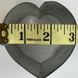 Puede incluir: Un cortador de galletas de metal en forma de corazón con acabado plateado. Una cinta métrica amarilla está colocada en el centro, mostrando un ancho de aproximadamente 7,6 centímetros. La cinta tiene números negros y el texto FIBER-GLASS.
