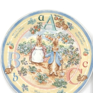 May include: A round ceramic plate featuring Peter Rabbit characters in a garden. The design includes the letters A, B, and C, along with illustrations of rabbits, fruits, and vegetables. Text includes 'Which fruit begins with b?' and 'Which vegetable begins with c?'