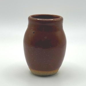 May include: A small, rounded ceramic vessel with a glossy, deep brown glaze covering the upper body and a contrasting, unglazed, textured beige base. The pot features a smooth, slightly flared rim and a simple, bulbous silhouette.