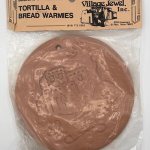 May include: A round, terracotta-colored ceramic tortilla and bread warmer in a clear plastic bag with a header card labeled Designs by Monique, Tortilla & Bread Warmies, and Village Jewel, Inc. The ceramic disc features an embossed desert scene.