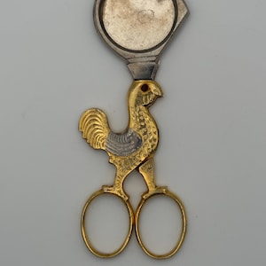 May include: A pair of gold-toned metal scissors featuring a rooster-shaped handle with intricate engraved feather details and two oval finger loops. The blades are topped with a circular, silver-toned metal magnifying glass attachment.