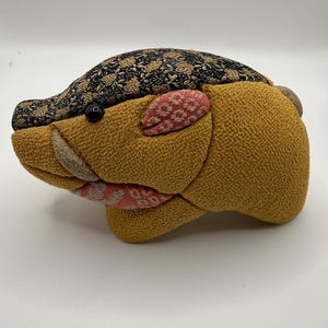 May include: A stuffed fabric animal shaped like a wild boar, featuring a textured mustard yellow body, a black and gold floral patterned back, and pink patterned accents on the ear and jaw. It has a small black bead eye and a tan tusk.