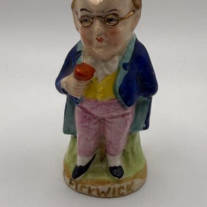 May include: A ceramic figurine of a character wearing gold-rimmed glasses, a blue coat, a yellow vest, and pink trousers. The figure holds a small red object in one hand and stands on a base with the word Pickwick inscribed in black lettering.
