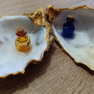 Può includere: Due conchiglie di ostrica aperte con bordi dipinti in oro e interni bianco perlaceo. Ogni conchiglia contiene una piccola bottiglia di vetro con tappo di sughero; una bottiglia è color ambra e l'altra è blu scuro, poggiate all'interno.