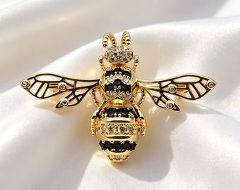 May include: A gold-toned bee brooch featuring black and clear crystal accents. The insect-shaped pin has detailed wings with small embedded stones, a head covered in clear crystals, and a striped abdomen alternating between black and clear stones.