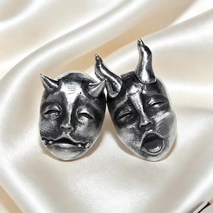 May include: Two silver-toned metal charms shaped like horned faces. The left charm features a closed-mouth expression with small horns, while the right charm depicts a face with an open mouth and larger, prominent horns, both with dark oxidation.