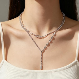 Può includere: Una collana a strati color argento caratterizzata da una combinazione di delicate catene a palline, maglie ovali allungate e una sezione di piccole maglie arrotondate a forma di ancora. Il design include una catena verticale con un pendente rettangolare.