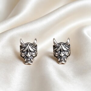 May include: A pair of silver-toned stud earrings featuring a detailed Hannya mask design. Each earring displays the distinct facial features of the mask, including prominent horns, a furrowed brow, and exposed fangs, finished with a dark oxidation.