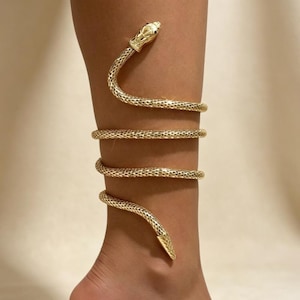Op de afbeelding: Een goudkleurige slangenarmband of beenwikkel met een getextureerd, schubachtig oppervlak. Het ontwerp spiraalt rond het ledemaat en eindigt bovenaan in een gedetailleerde slangenkop met kleine zwarte ogen en onderaan in een toelopende staart.