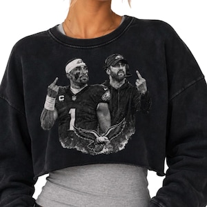 Könnte beinhalten: Ein schwarzes, säuregewaschenes bauchfreies Sweatshirt mit einem Grafikdruck eines Footballspielers und eines Trainers, die beide den Mittelfinger zeigen. Das Design enthält ein Adler-Emblem und die Nummer eins auf dem Trikot des Spielers.