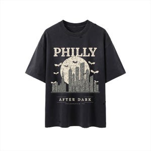 Könnte beinhalten: Ein anthrazitfarbenes Kurzarm-T-Shirt mit Used-Optik. Die Grafik zeigt einen weißen Mond, eine graue Skyline und fliegende Fledermäuse. Der Text lautet PHILLY, AFTER DARK und PHILADELPHIA, PA in einer kräftigen, verwitterten weißen Schrift.