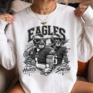 Könnte beinhalten: Ein weißes Sweatshirt mit Rundhalsausschnitt und einem schwarz-weißen Grafikdruck der Philadelphia Eagles-Spieler Jalen Hurts und DeVonta Smith in ihren Uniformen. Der Text lautet EAGLES, JALEN HURTS, 1, DEVONTA SMITH und 6, mit einem Adler-Logo.