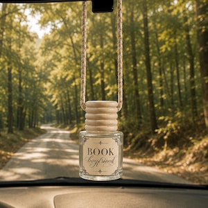 Könnte beinhalten: Eine durchsichtige Auto-Duftflasche aus Glas mit einem hellen Holzdeckel und einer geflochtenen Kordel zum Aufhängen. Das Etikett hat einen dekorativen Rand und den schwarzen Schriftzug "BOOK boyfriend". Eine kleine Glasvase mit Trockenblumen ist in der Nähe befestigt.