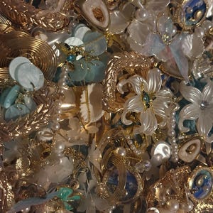 May include: A collection of gold-toned jewelry pieces featuring white floral accents, faux pearls, and light blue circular elements. The assortment includes various earrings and brooches with intricate metal textures and clear crystals.