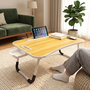 May include: A portable laptop desk with a light wood grain surface, white frame, and black leg grips. It features a built-in tablet slot, a small pull-out side drawer, and a handle cutout. The desk is positioned for use with a tablet and books.