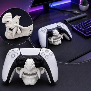 May include: A white gaming controller stand shaped like a stylized samurai mask with prominent fangs and intricate facial details. The stand is designed to hold a white and black wireless game controller securely in place on a flat surface.