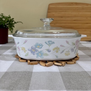 Puede incluir: Una fuente de horno ovalada de color blanco con tapa de cristal transparente y asas laterales integradas. El exterior presenta un delicado estampado floral con flores de color azul claro, rosa y amarillo y hojas verdes que rodean el recipiente.