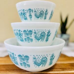 Peut inclure: Un ensemble de trois bols mélangeurs en verre opalin blanc empilés verticalement. Chaque bol présente un motif répétitif bleu sarcelle composé d'une personne tenant un râteau, de gerbes de blé, d'un coq et d'épis de maïs sur le bord extérieur.