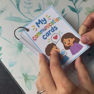 Può includere: Un mazzo di carte di comunicazione rettangolari rilegate da un anello in metallo argentato. La carta frontale presenta il testo My communication cards in lettere colorate, un'icona a forma di cuore rosso e un'illustrazione di due persone.