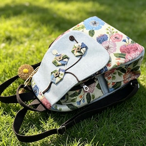 Op de afbeelding: Een rugzak met een bloemenpatroon van blauwe en roze bloemen op een crèmekleurige achtergrond. De voorflap is lichtblauw met drie stoffen zeshoekige versieringen en donkerbruine banden. Een gele pompon hangt aan de zijkant.