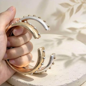 May include: Two open-ended cuff bracelets, one in a polished gold tone and one in a silver tone, each featuring a series of small, evenly spaced spherical studs along the outer edge of the curved metal band.
