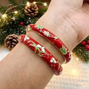 Puede incluir: Dos pulseras rígidas de color rojo con un patrón floral de esmalte en blanco y verde. Las pulseras tienen un acabado liso y brillante con bordes de metal dorado. El diseño floral incluye pétalos blancos y hojas verdes.