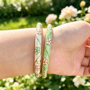 Könnte beinhalten: Zwei Armreifen mit Blumenmotiven. Ein Armreif ist cremefarben mit rosa und hellgrünen Blüten, der andere ist hellgrün mit rosafarbenen Blüten. Beide Armreifen sind mit feinen, goldfarbenen, metallischen Rankenmustern verziert.