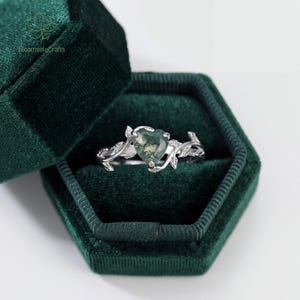 May include: A silver ring featuring a heart-shaped moss agate gemstone set in a band designed with intricate metal leaves and vines. The ring is displayed inside a dark green velvet hexagonal box with the text BloomvaleCrafts printed on the lid.