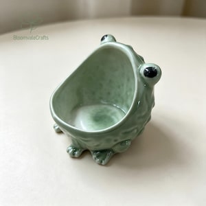 Peut inclure: Un bol en céramique vert clair en forme de grenouille avec une finition brillante. Le bol présente un extérieur texturé, deux yeux proéminents sur le bord supérieur et un intérieur lisse et concave. Le logo BloomvaleCrafts est visible.