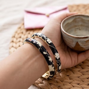 Pode incluir: Dois braceletes pretos com um padrão floral branco e detalhes delicados de vinhas. Os braceletes têm um acabamento liso e brilhante, com formato circular, exibindo um design botânico repetitivo em toda a superfície de cada peça.