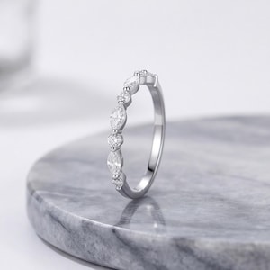 May include: A silver-toned ring featuring a series of alternating marquise-cut and round-cut clear stones set along the top half of the band. The metal band has a polished finish and a slender profile, showcasing a delicate and symmetrical design.