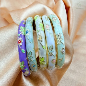 May include: A set of five bangle bracelets featuring smooth enamel surfaces in shades of purple, light blue, white, and pale green. Each bracelet is decorated with delicate gold-toned floral and leaf patterns that wrap around the entire circumference.