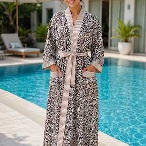 May include: A full-length robe featuring a brown and black leopard print pattern. The garment includes a beige and white striped trim along the front opening, sleeve cuffs, and two front pockets. A matching fabric belt ties at the waist.