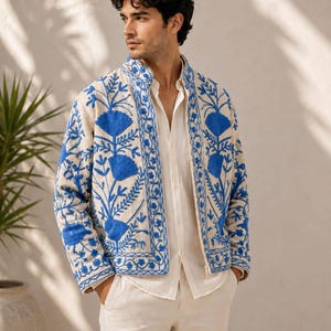 May include: A beige jacket featuring vibrant blue floral embroidery along the front panels and collar. The jacket has a structured stand-up collar, long sleeves, and a full-length front zipper closure, worn over a plain light beige shirt.