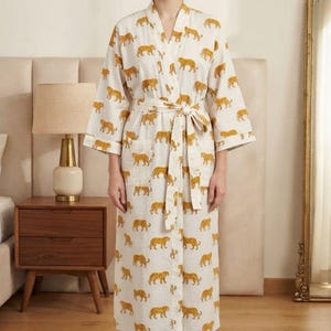 May include: A long-sleeved, calf-length robe in an off-white colour featuring a repeating pattern of small, mustard-yellow tigers. The robe includes a matching fabric tie belt at the waist, wide sleeves, and two front patch pockets on the lower section.