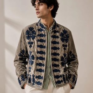 Può includere: Una giacca in velluto color tortora con colletto alla coreana e ricami floreali blu scuro sui pannelli frontali e sui polsini. La giacca è indossata sopra una camicia a righe verticali verde chiaro e bianco e pantaloni color bianco sporco.