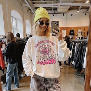 May include: A white long-sleeve sweatshirt featuring a graphic print with the text "ACADEMIC SUPPORT" and "DREAM Team" in pink and brown, accompanied by a central illustration of daisies tied with a pink ribbon and small pink stars.