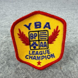 May include: A yellow shield-shaped embroidered patch with a red border. It features blue text reading YBA LEAGUE CHAMPION, a central square divided into four quadrants with the letters BP, AA, and symbols, and two red leaf motifs.