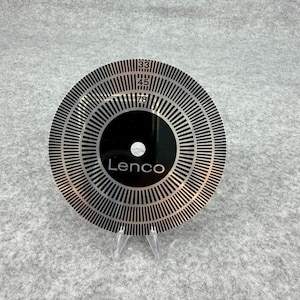May include: A circular black and silver Lenco strobe disc featuring concentric rings of vertical lines and numerical markings for 33, 45, and 78 RPM speeds. The disc has a central hole and a smooth, reflective finish with clear, high-contrast graphics.