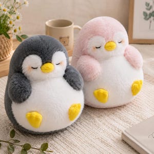 Op de afbeelding: Twee pluche pinguïnknuffels met zachte, ronde lichamen. De ene pinguïn is donkergrijs en wit, terwijl de andere lichtroze en wit is. Beide hebben gesloten ogen, kleine gele snavels en gele voetjes, uitgevoerd in een zachte, pluizige stof.
