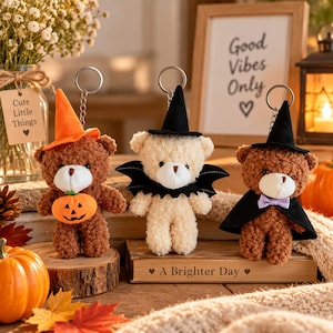 Może przedstawiać: Trzy pluszowe breloki w kształcie misiów z motywami halloweenowymi. Pierwszy jest brązowy z pomarańczowym kapeluszem czarownicy i dynią. Drugi jest kremowy z czarnym kapeluszem czarownicy i skrzydłami nietoperza. Trzeci jest brązowy z czarnym kapeluszem.