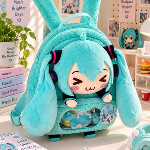 May include: A plush turquoise backpack featuring a soft, fuzzy texture and long rabbit-like ears. The front displays a cartoon character face with teal hair, black and pink headphones, and a clear window pocket containing small items and a circular charm.