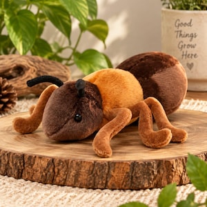 Op de afbeelding: Een pluche mierenknuffel met een donkerbruine kop en achterlijf en een goudbruin borststuk. Het knuffeldier heeft twee zwarte voelsprieten, zes poten en kleine zwarte oogjes. Het speelgoed ligt op een ronde houten schijf met een textuur.