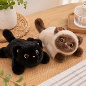 Peut inclure: Deux jouets en peluche en forme de chat en position allongée. Un chat est entièrement noir avec de grands yeux ambrés et un petit nez rose. Le second chat a un pelage crème sur le corps avec un visage, des oreilles et une queue brun foncé.