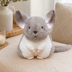 Könnte beinhalten: Ein Chinchilla-Stofftier mit weichem grauem Fell an Kopf und Rücken, einem weißen Bauch und großen, aufrechten Ohren mit glattem Innenfutter. Das Plüschtier hat kleine schwarze Augen, eine dunkle Nase, feine Schnurrhaare und hellrosa Pfoten.