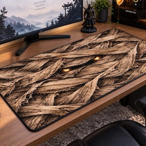 May include: A rectangular desk mat featuring a detailed graphic print of layered, textured tan and brown mummy bandages. Two glowing, bright yellow eyes are visible peering out from between the fabric wrappings in the center of the mat.