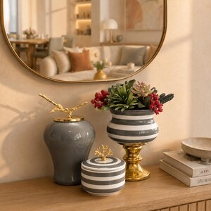 May include: A collection of three decorative ceramic vessels: a tall glossy grey jar with a gold branch lid, a small round grey and white striped jar with a gold coral lid, and a grey and white striped pedestal bowl filled with faux succulents.