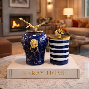 Peut inclure: Deux bocaux décoratifs posés sur un livre blanc marqué FERAY HOME. Le bocal de gauche est bleu foncé avec des veines de marbre doré, un heurtoir de porte en forme de tête de lion dorée et un couvercle branche doré. Le droit a des rayures bleu marine.