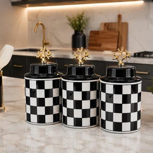 Puede incluir: Tres botes cilíndricos de cerámica con un patrón de cuadros blancos y negros. Cada bote tiene una tapa negra a juego rematada con un remate decorativo de metal dorado con forma de estructura de coral ramificado.